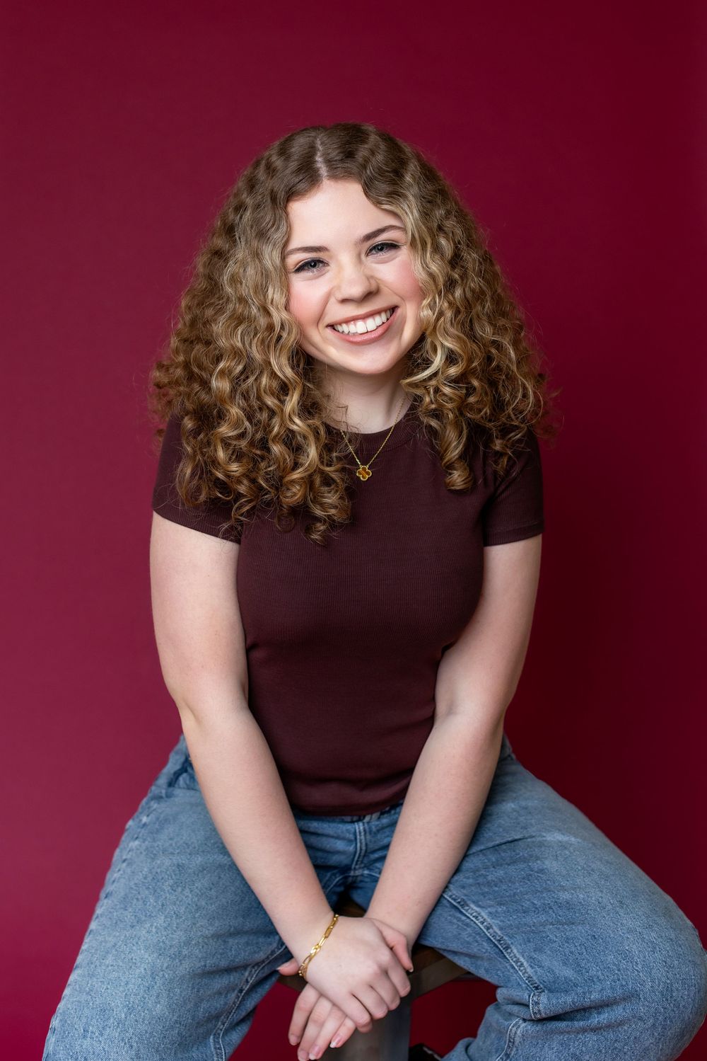 Smiling young woman with curly hair wearing a maroon top and jeans, seated against a red background.
