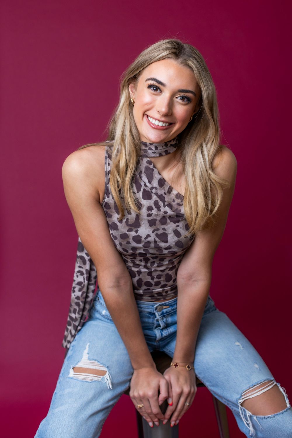 Smiling blonde woman in leopard print top and ripped jeans poses on stool against crimson background.