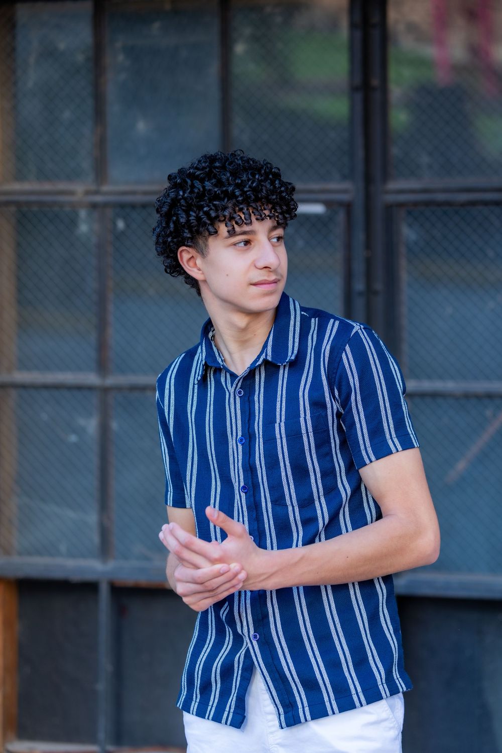 Young man with curly black hair wearing a blue striped short-sleeve shirt and white pants standing outdoors.