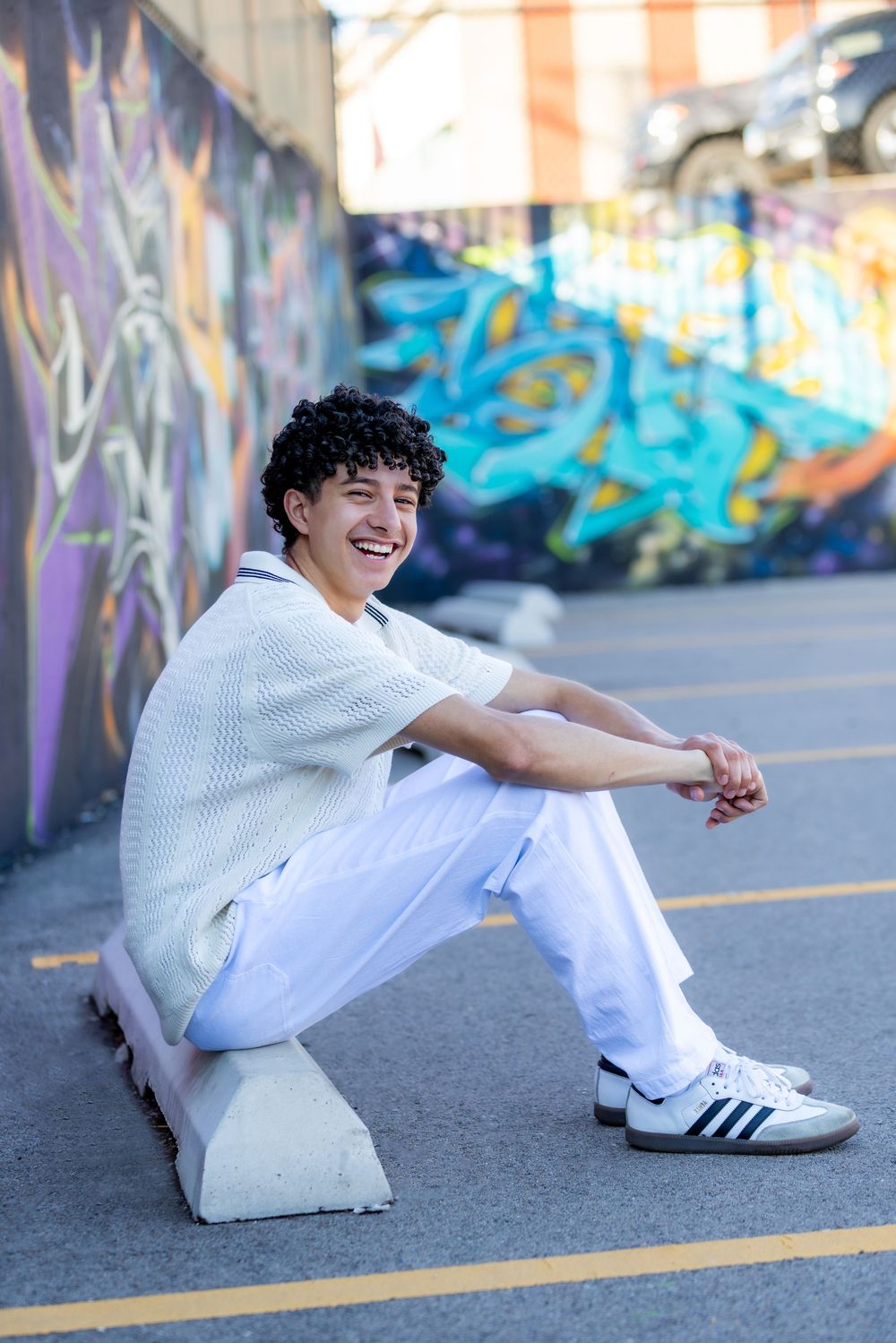 Smiling young man in white outfit sitting on concrete block in front of colorful graffiti wall.
