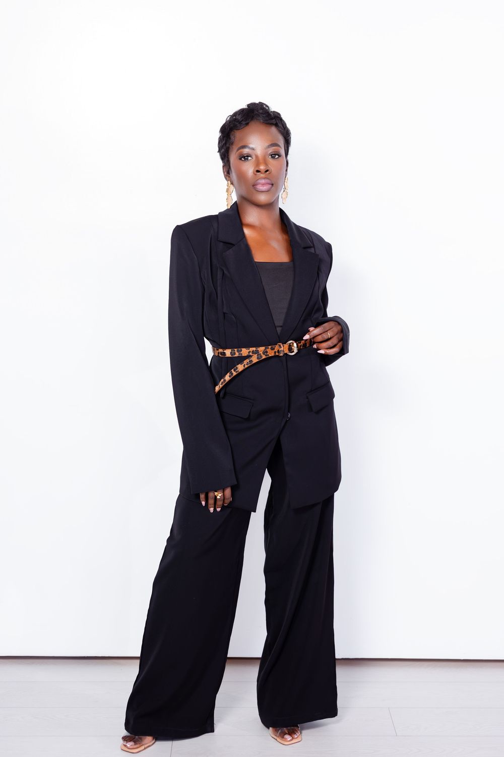 Fashion model in black power suit and wide leg pants poses confidently against white background.