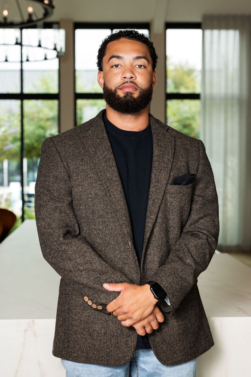 Professional wearing a black turtleneck and tweed blazer poses in a modern office setting with natural lighting.