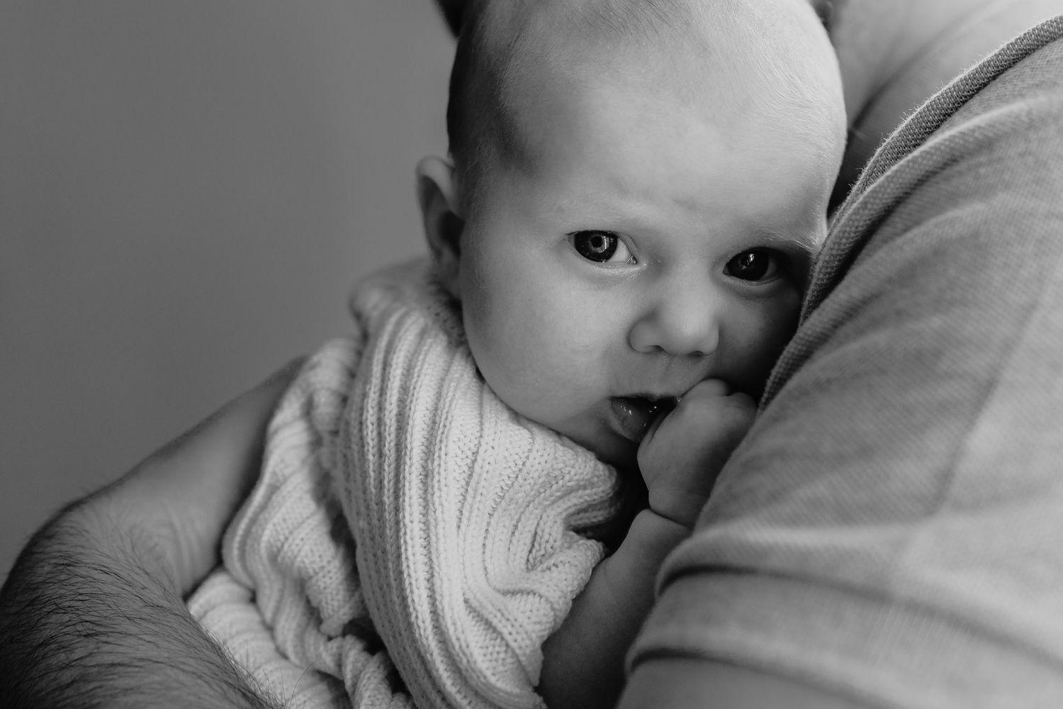Black and white portrait of a baby in a sweater leaning against an adult's arm.