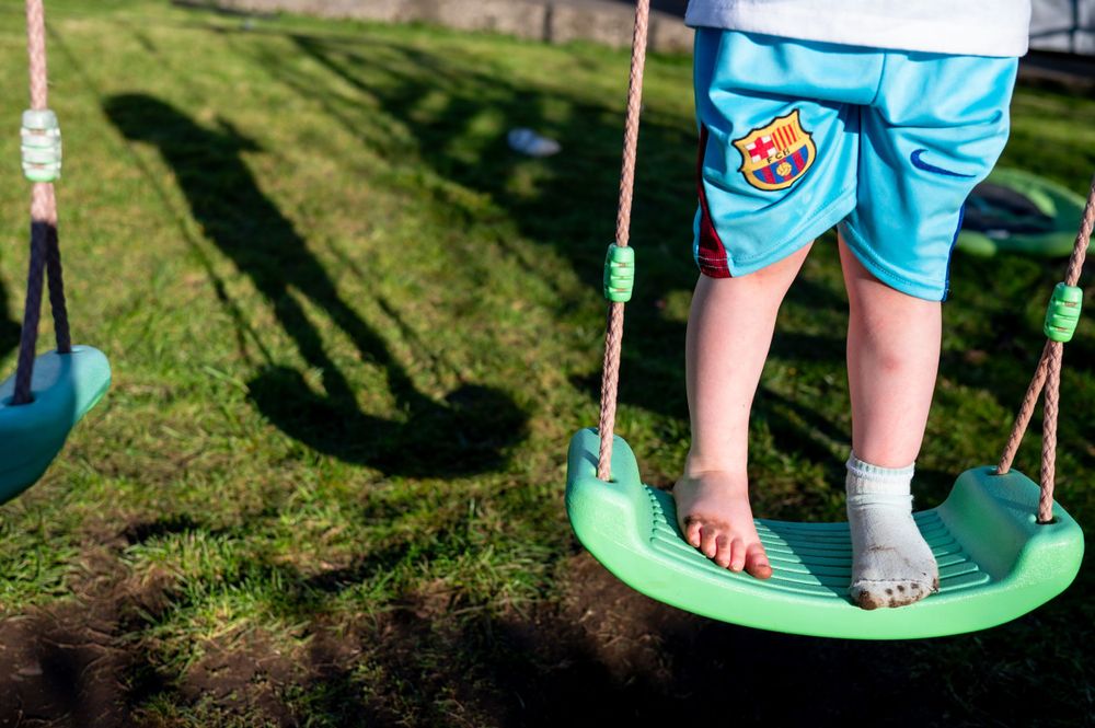Child wearing turquoise shorts standing barefoot on a green disc swing in a sunny backyard.