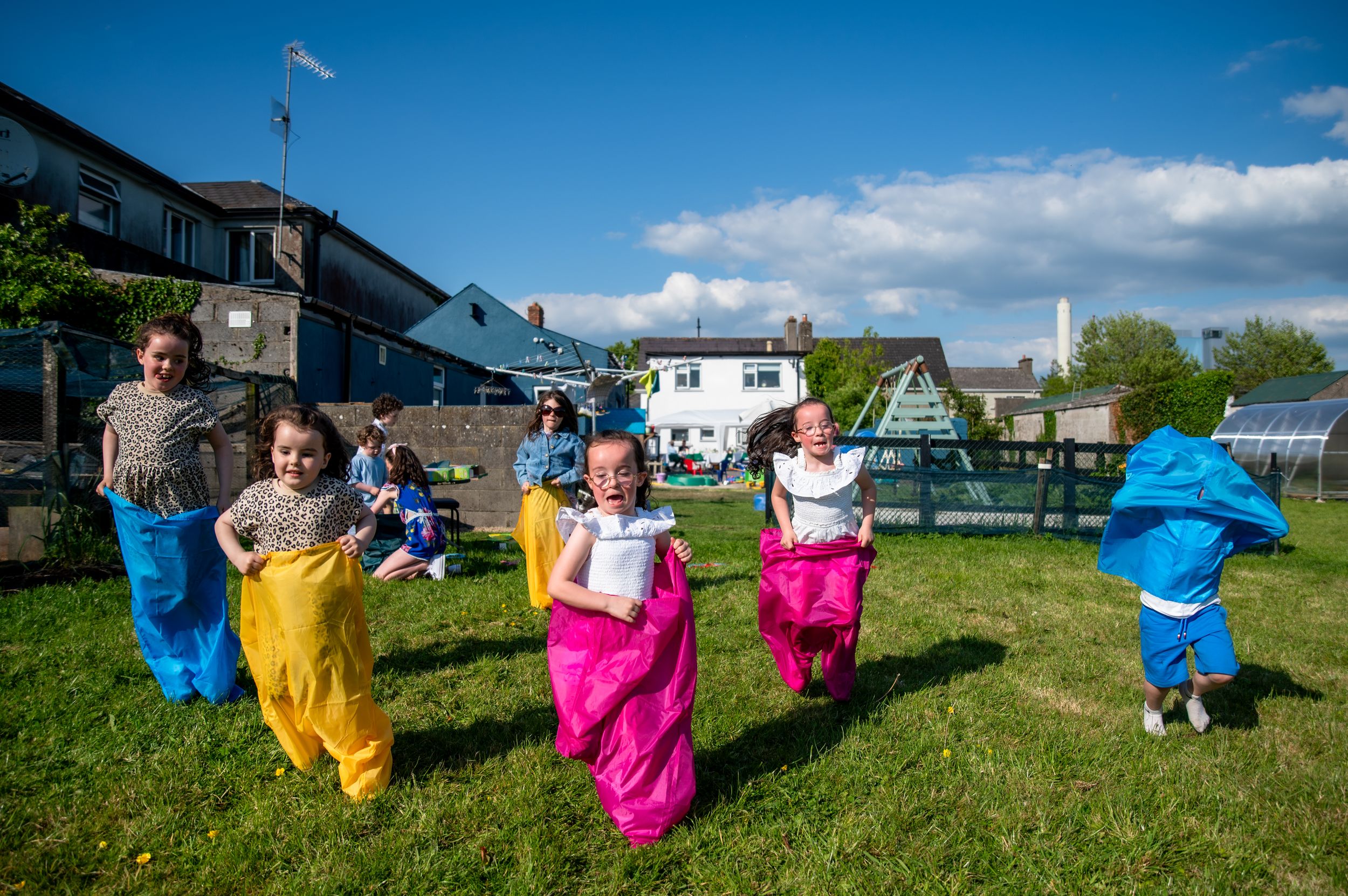 People enjoying a sack race competition on a sunny day in a grassy backyard with buildings in the background.