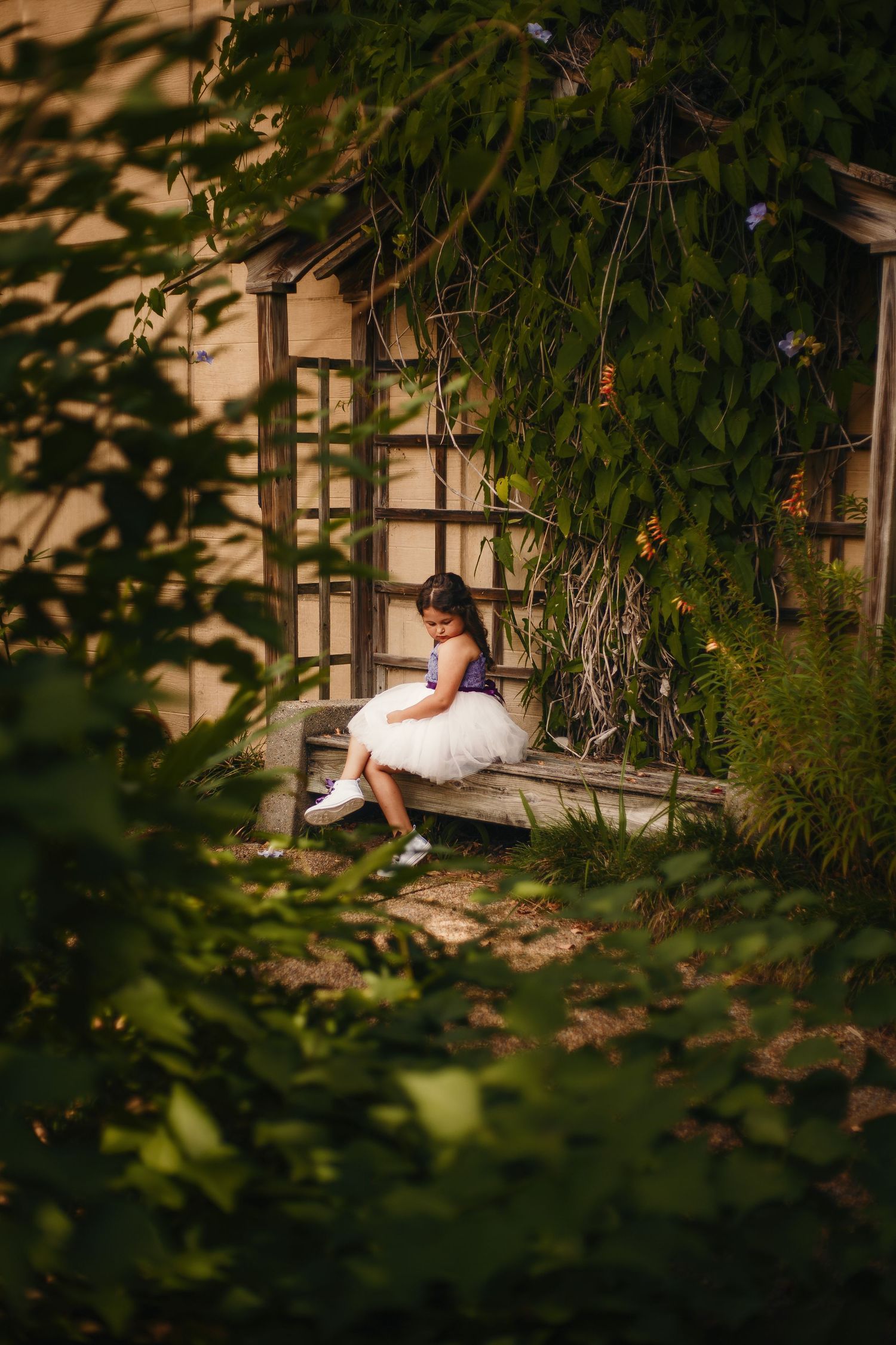 Little girl in white tutu dress sitting on steps surrounded by lush garden greenery.