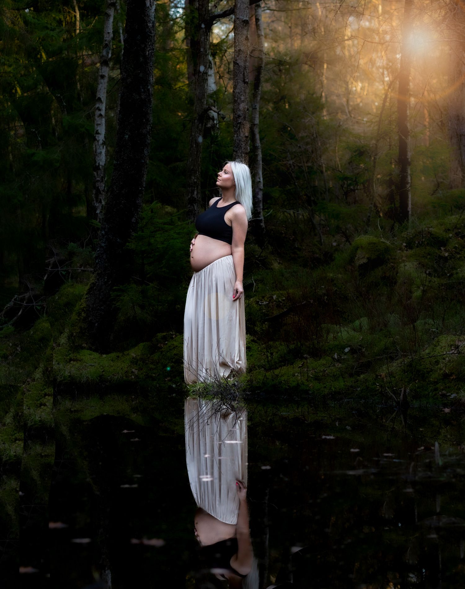 A reflection creates a mirror image of an expectant mother in a long dress standing by a tranquil forest pond at sunset.