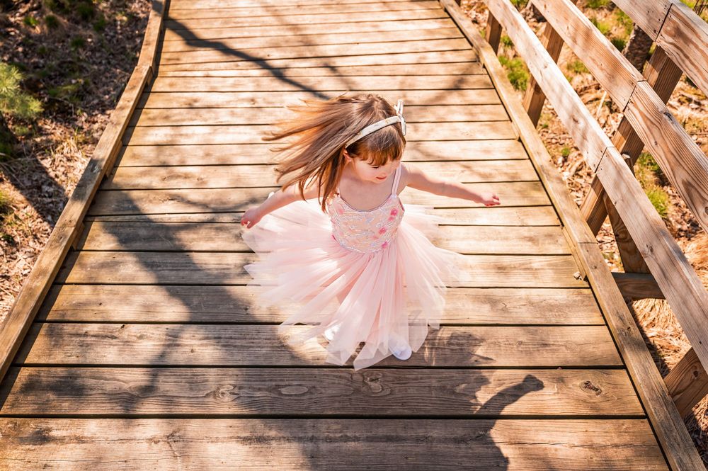 A small figure in a pink tulle dress twirls on a wooden boardwalk path surrounded by autumn foliage.