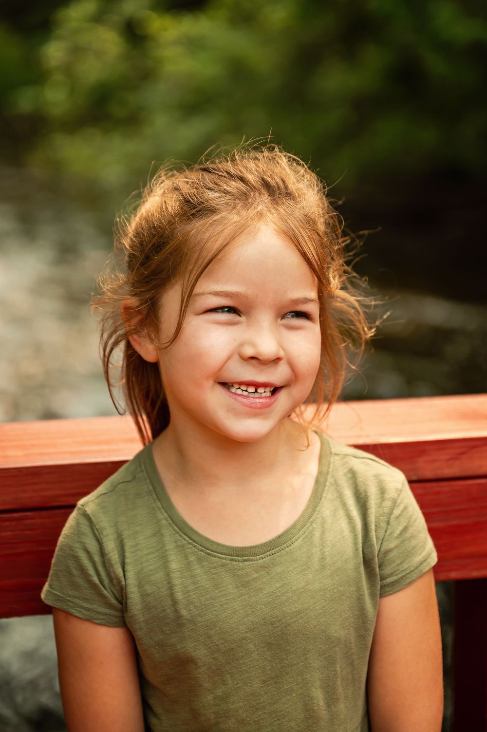 A child in a green t-shirt sits on a red bench outdoors, smiling with a joyful expression.