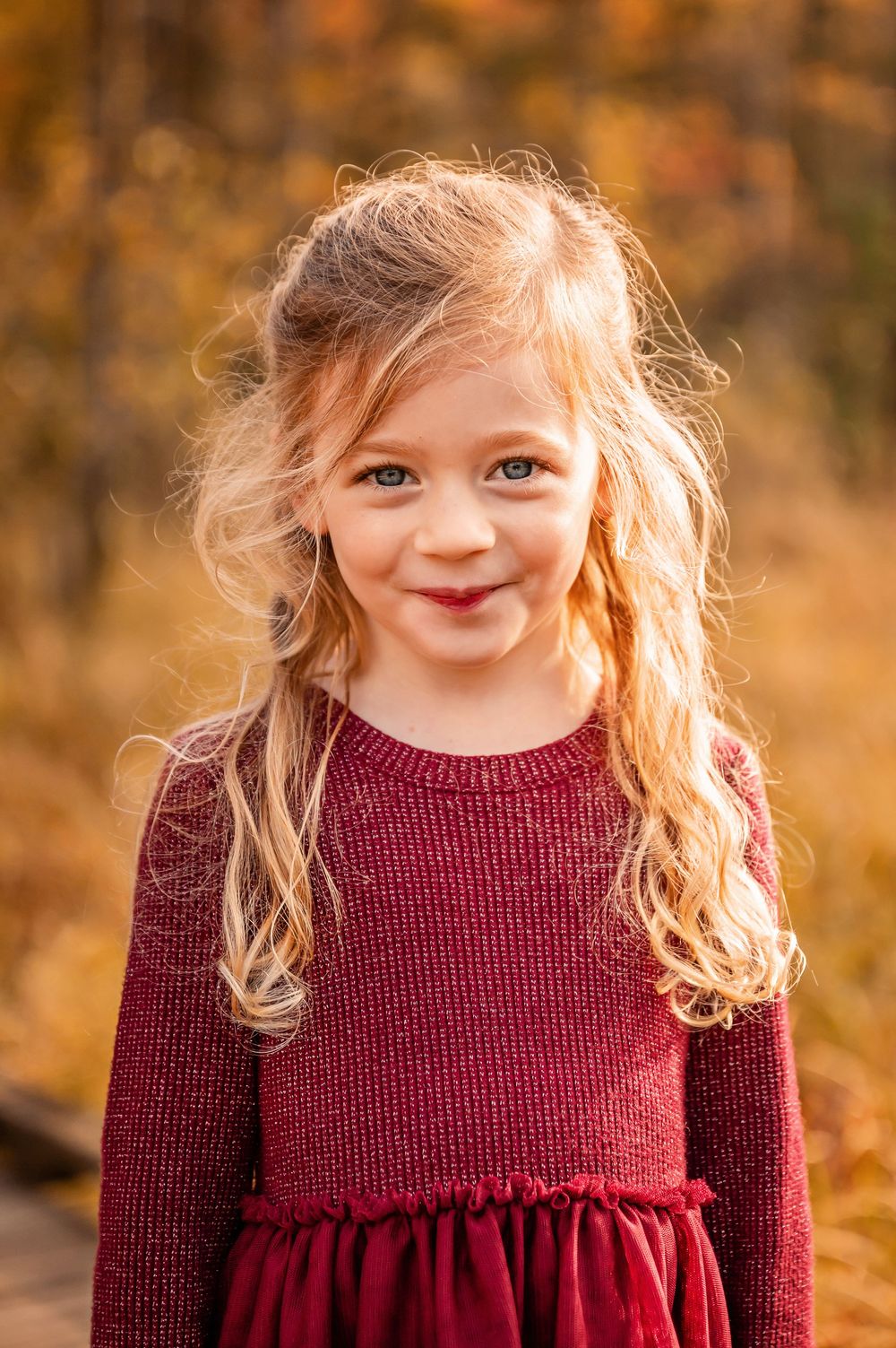 Portrait of a young child in a burgundy sweater dress smiling warmly against an autumn background with golden bokeh.