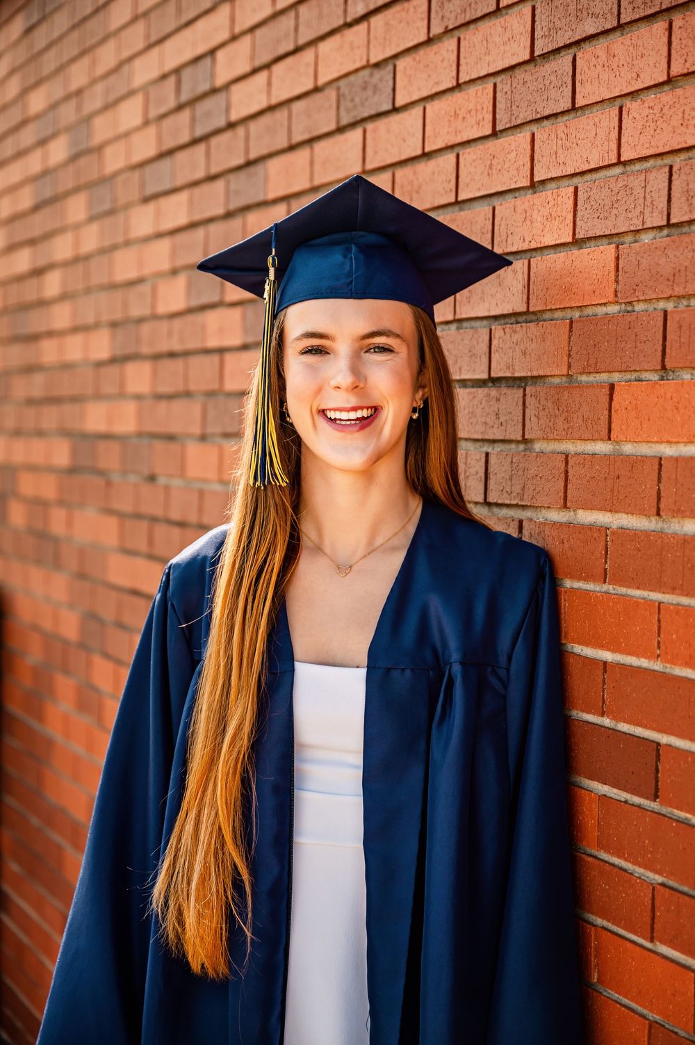 Graduate in navy blue cap and gown smiles against red brick wall in graduation photo series.