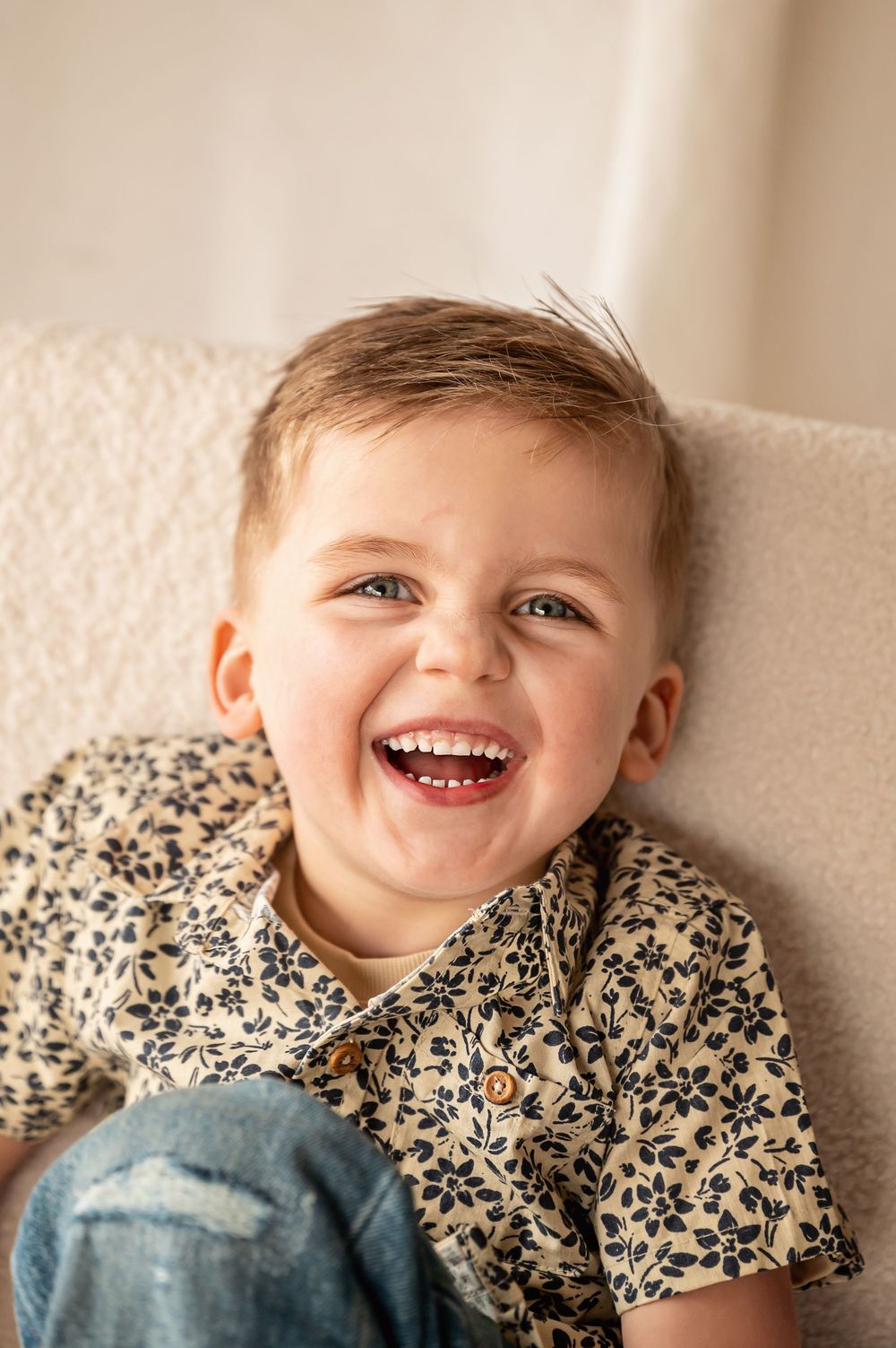A joyful child wearing a floral print shirt laughs while leaning against a cream-colored wall.
