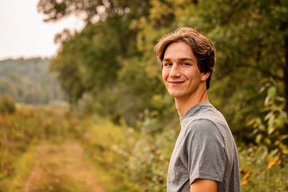 A young person in a gray t-shirt smiles while standing in an orchard on a sunny autumn day.