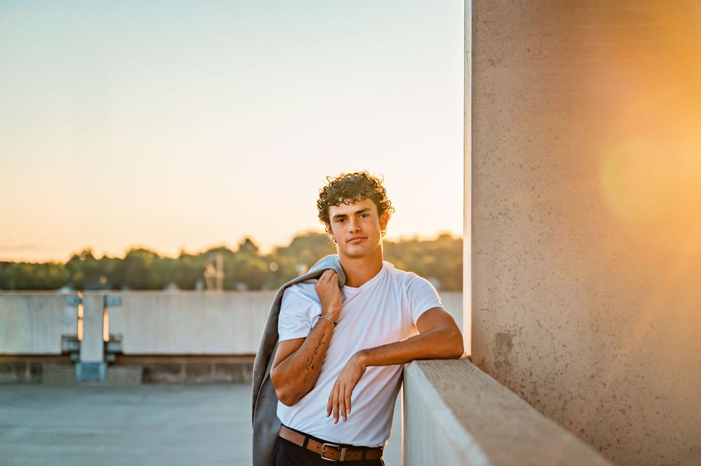 Young man leaning against wall during golden hour sunset with urban backdrop.