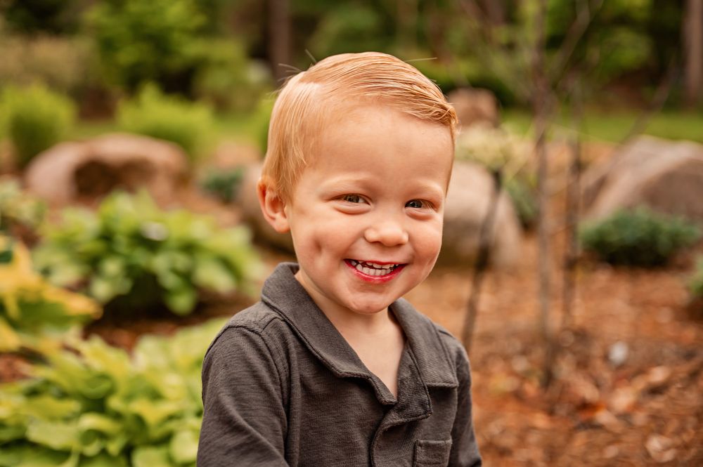A smiling child plays outdoors in a natural garden setting surrounded by green plants and brown mulch.