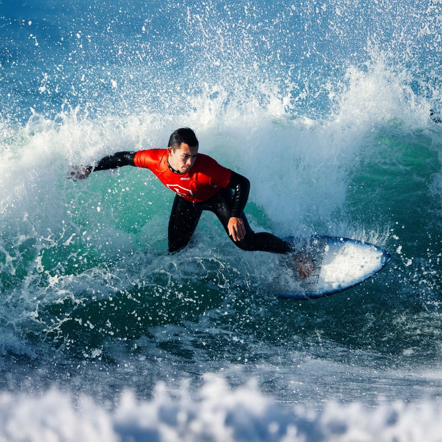 High school surfer in a red Orange Lutheran jersey catching a wave during a competition, surrounded by water spray.