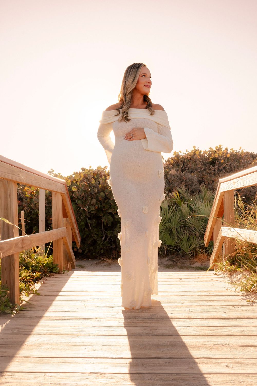 Pregnant woman in white off-shoulder gown standing on wooden boardwalk at sunset.