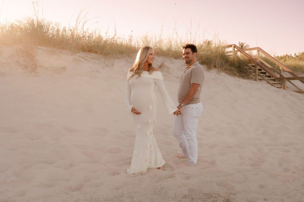 Expecting couple standing on sandy beach dunes at golden hour sunset together.