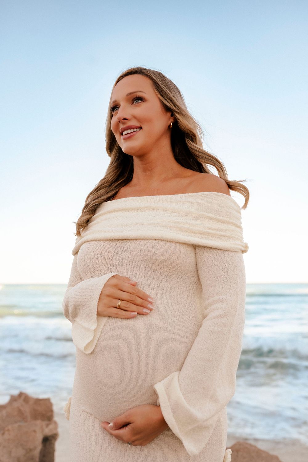 Smiling pregnant woman in off-shoulder cream dress cradling baby bump at beach.