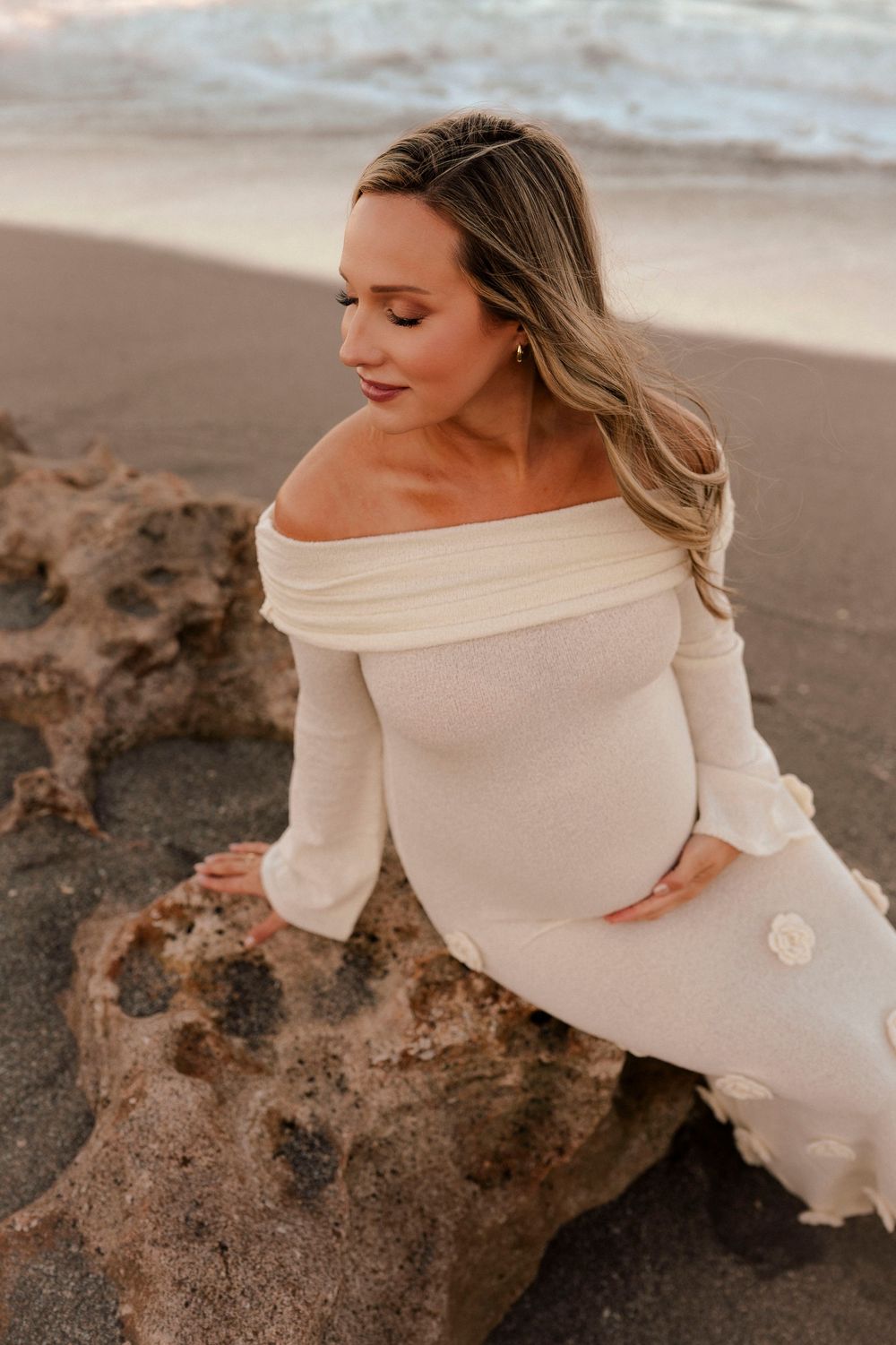 Pregnant woman in white off-shoulder dress sitting on beach rock at sunset.