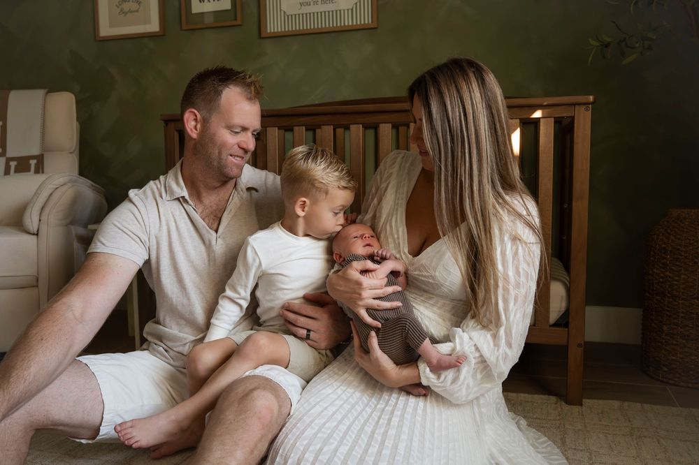 Happy family of four sitting on nursery floor with toddler and newborn baby.