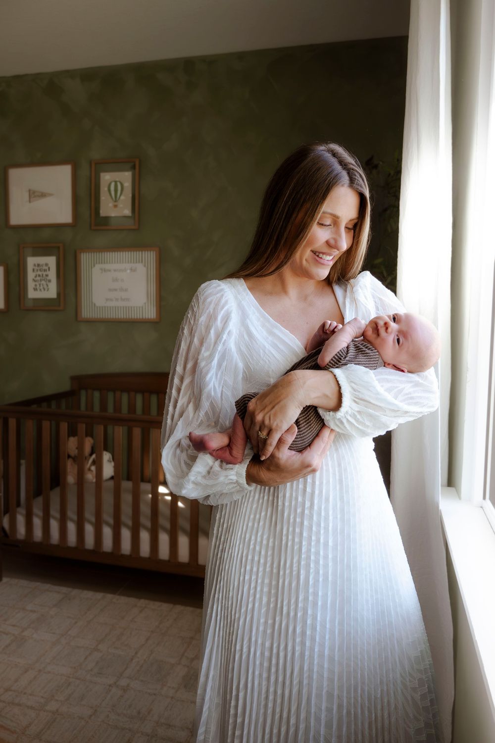 Smiling brunette mother in white dress holding newborn baby by nursery window.