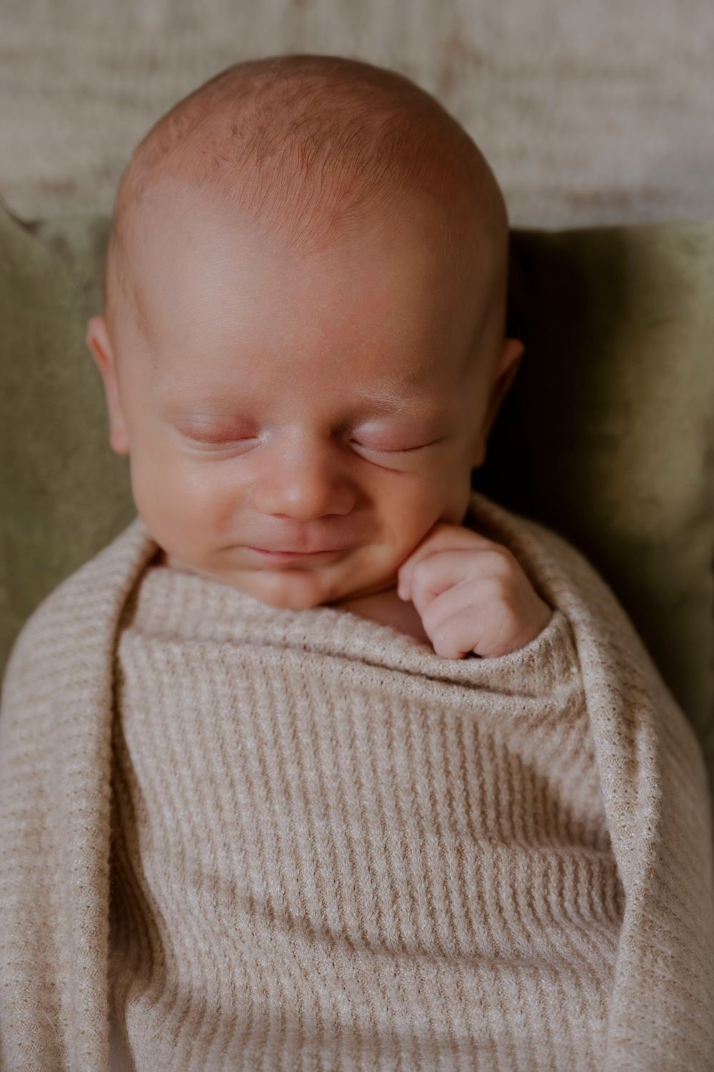 Sleeping newborn baby wrapped snugly in beige knit swaddle with tiny fist raised.