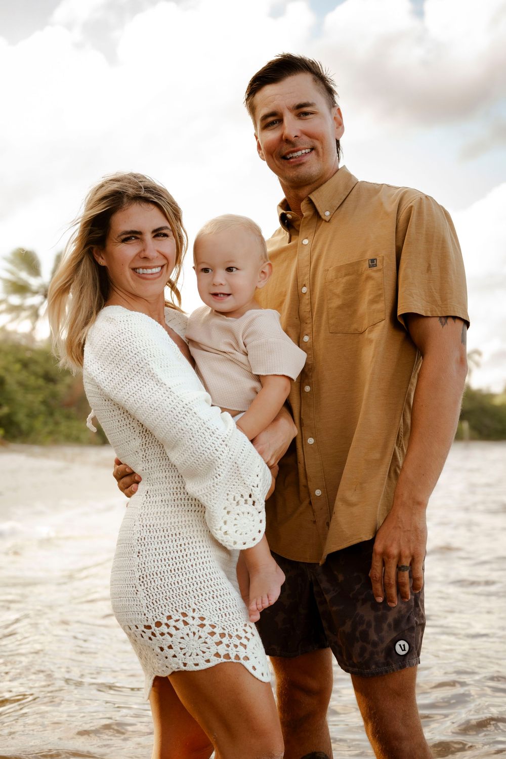 Smiling family of three with baby on tropical beach in golden sunset light.