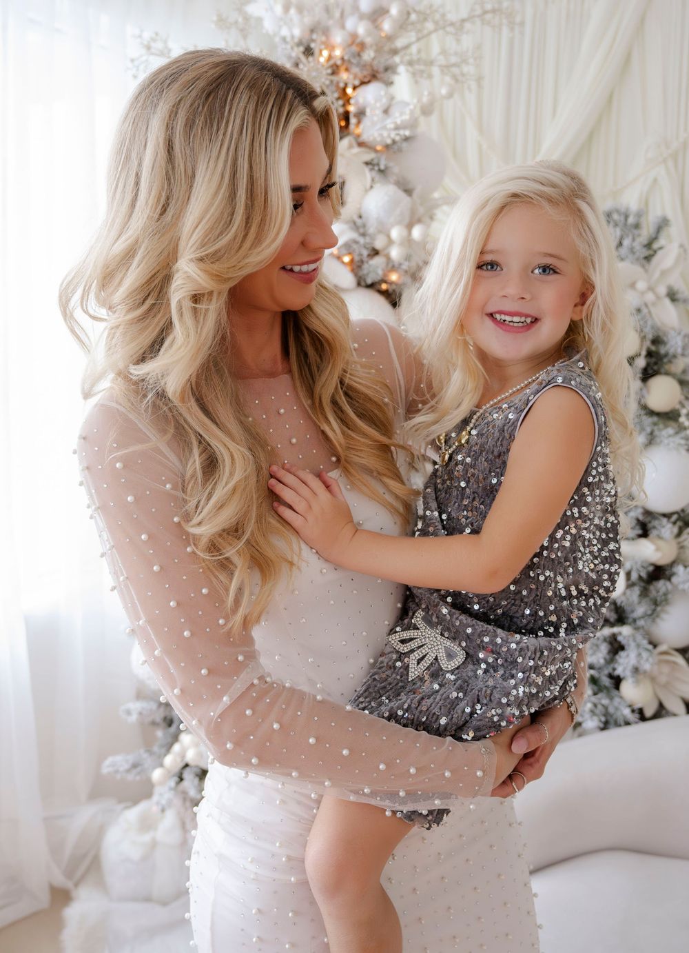 Blonde mother holding smiling daughter in sparkly dress by Christmas tree.