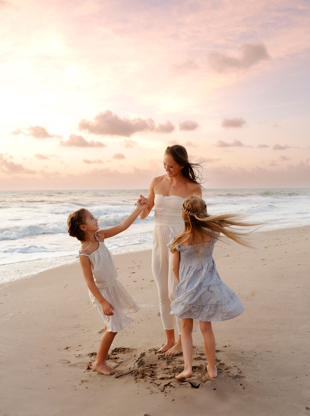 Mother dancing with two daughters on beach at pink sunset, wearing white dresses.