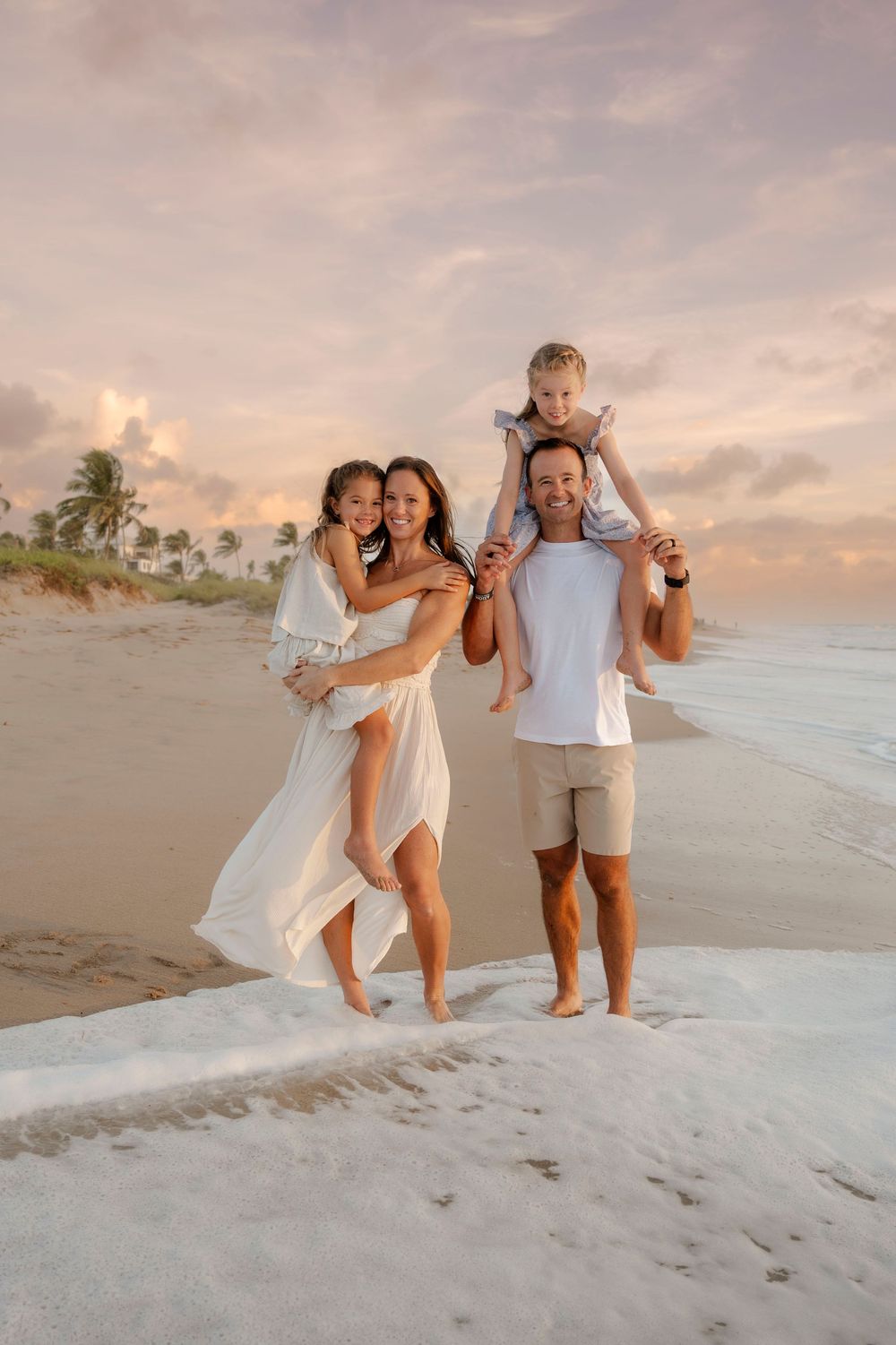 Happy family of four posing on tropical beach at sunset, parents holding children.