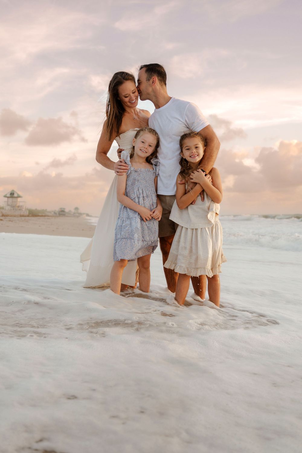 Loving family of four embracing on beach at sunset, parents kissing and hugging daughters.Palm Beach family photographer