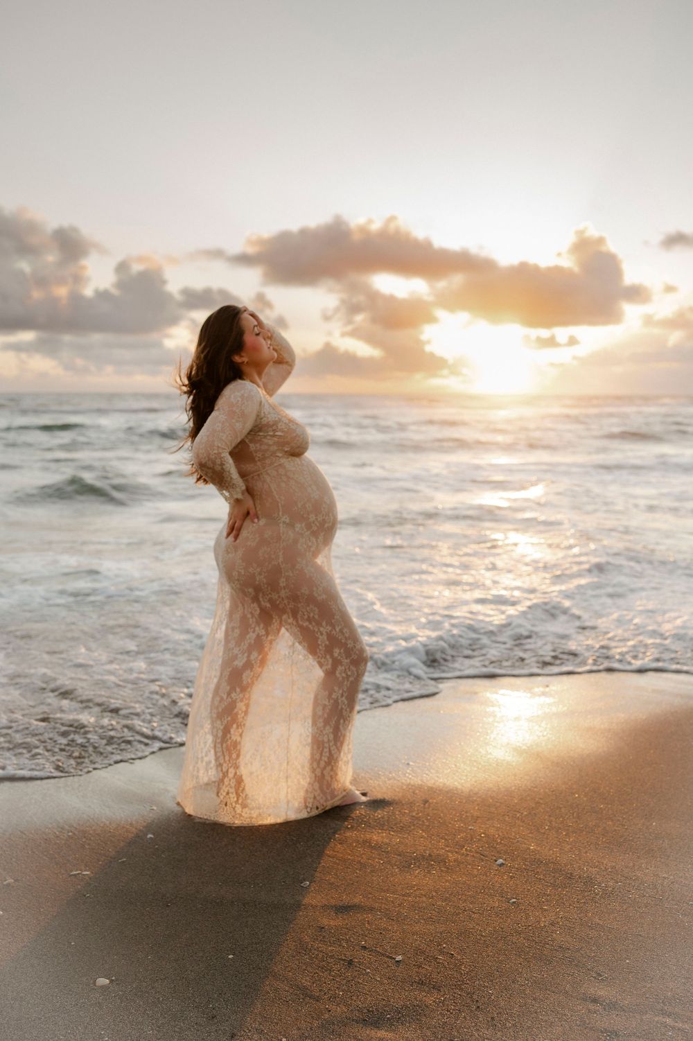 Pregnant woman in sheer lace dress standing on beach shore at golden sunset.