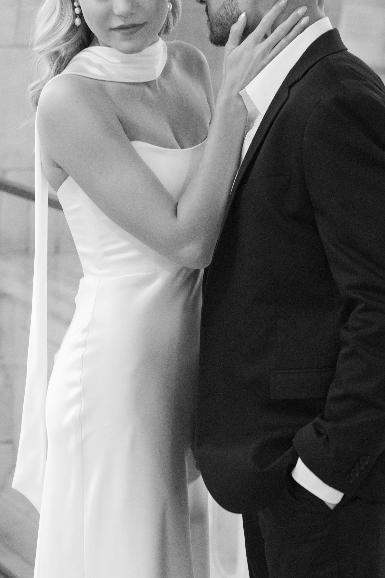 Black and white close-up of bride in sleek white halter gown embracing groom in black tuxedo tenderly.