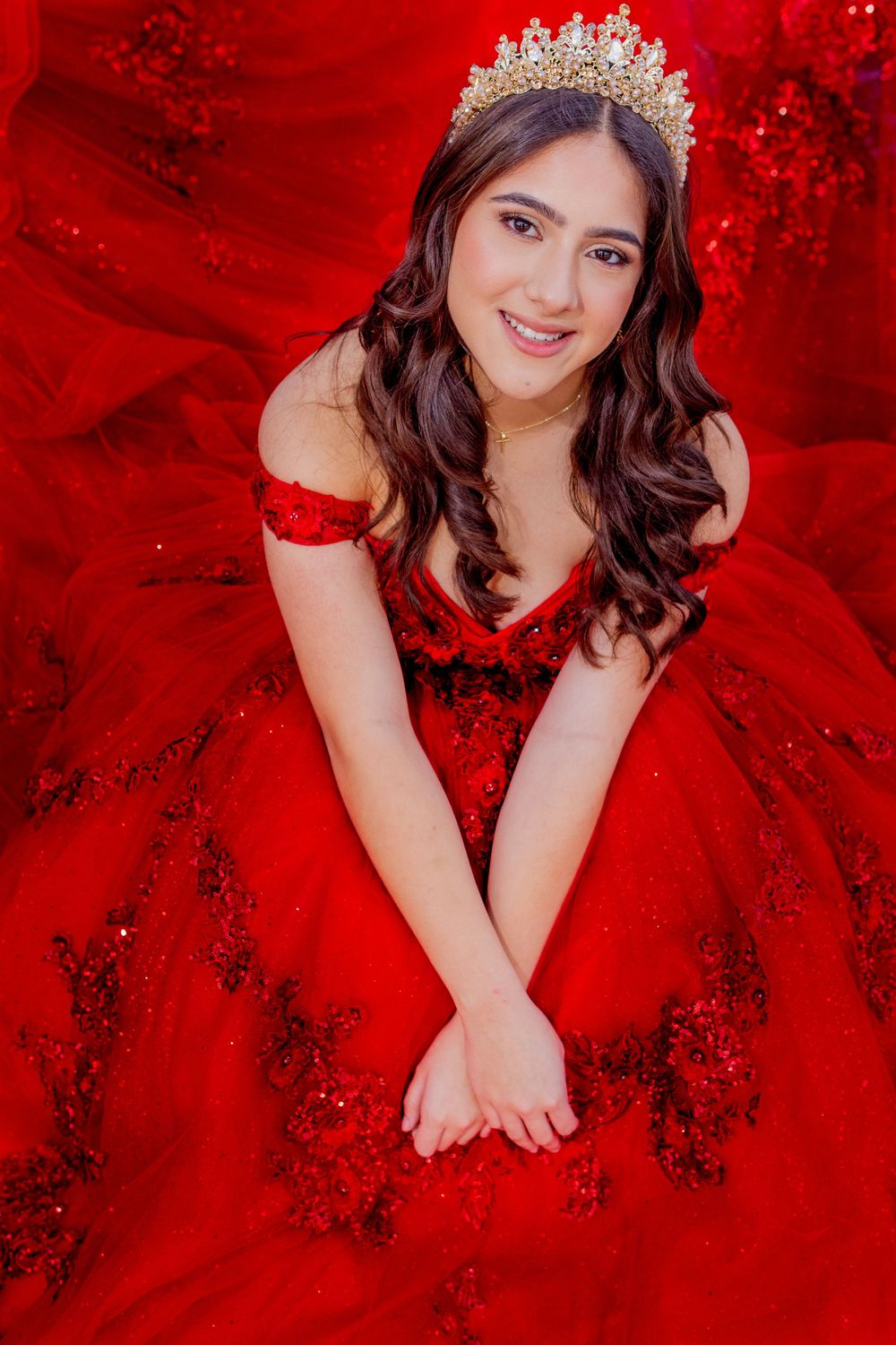 A sparkling red ballgown with crystal embellishments and tiara creates a dramatic portrait against red backdrop.