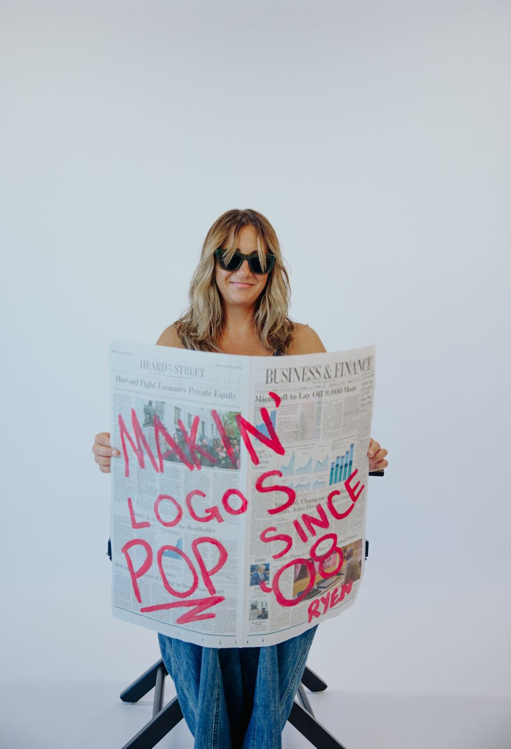 Woman holding newspaper with 'Makin Logos Pop Since 08' written in pink marker, wearing sunglasses.
