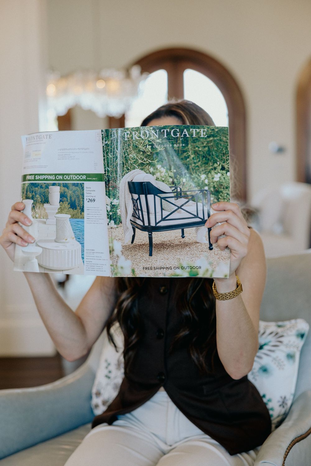 Woman sitting on a couch holding a Frontgate outdoor furniture catalog in a stylish home.