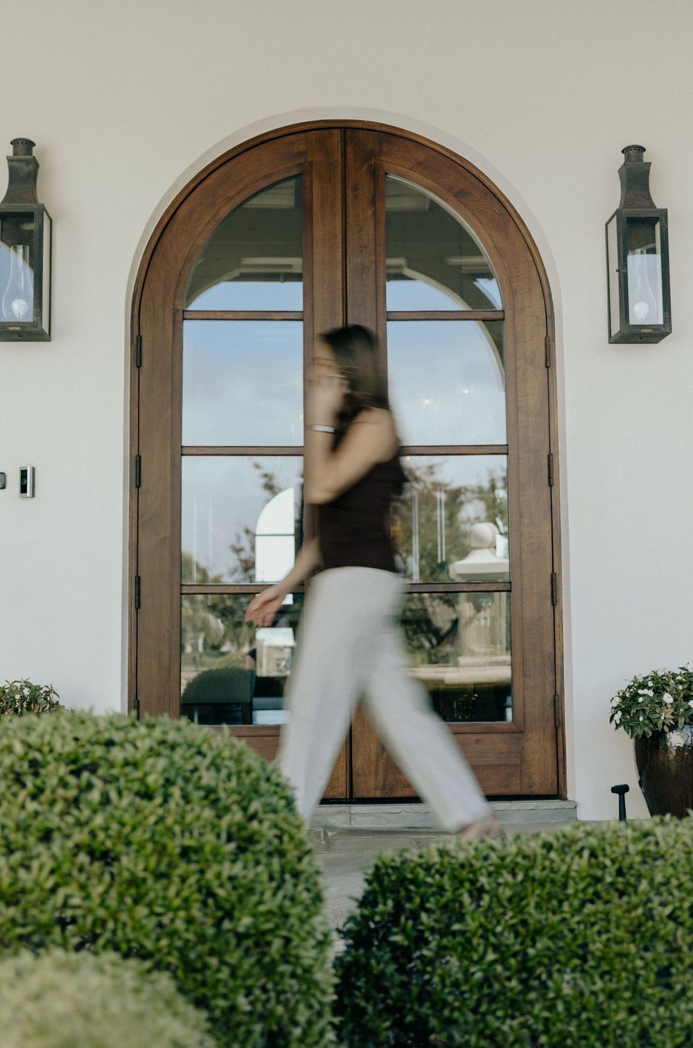 Woman in white pants walking past elegant arched wooden double doors with lush landscaping.