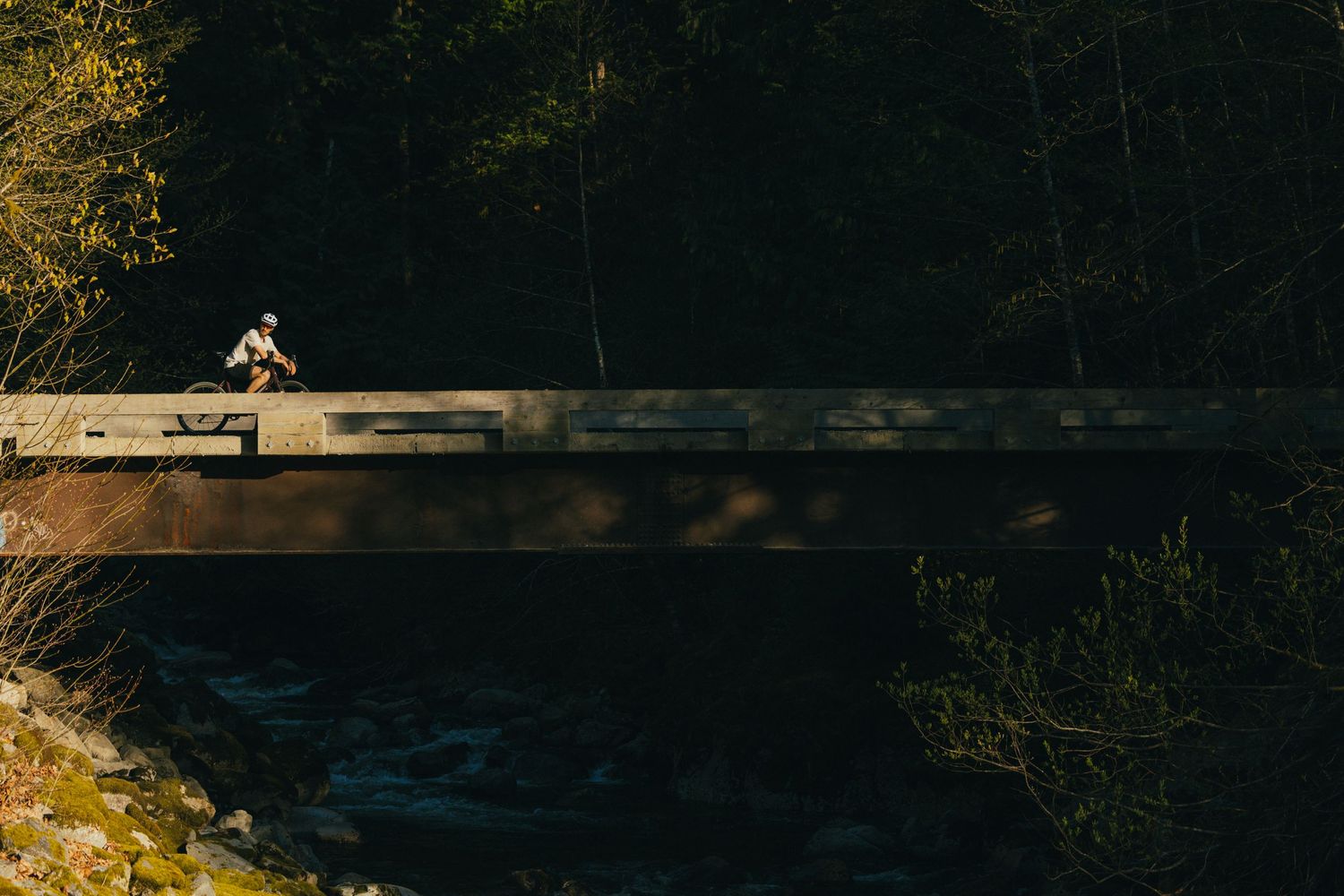 Distant cyclist silhouetted on wooden bridge over rushing stream in forest setting.
