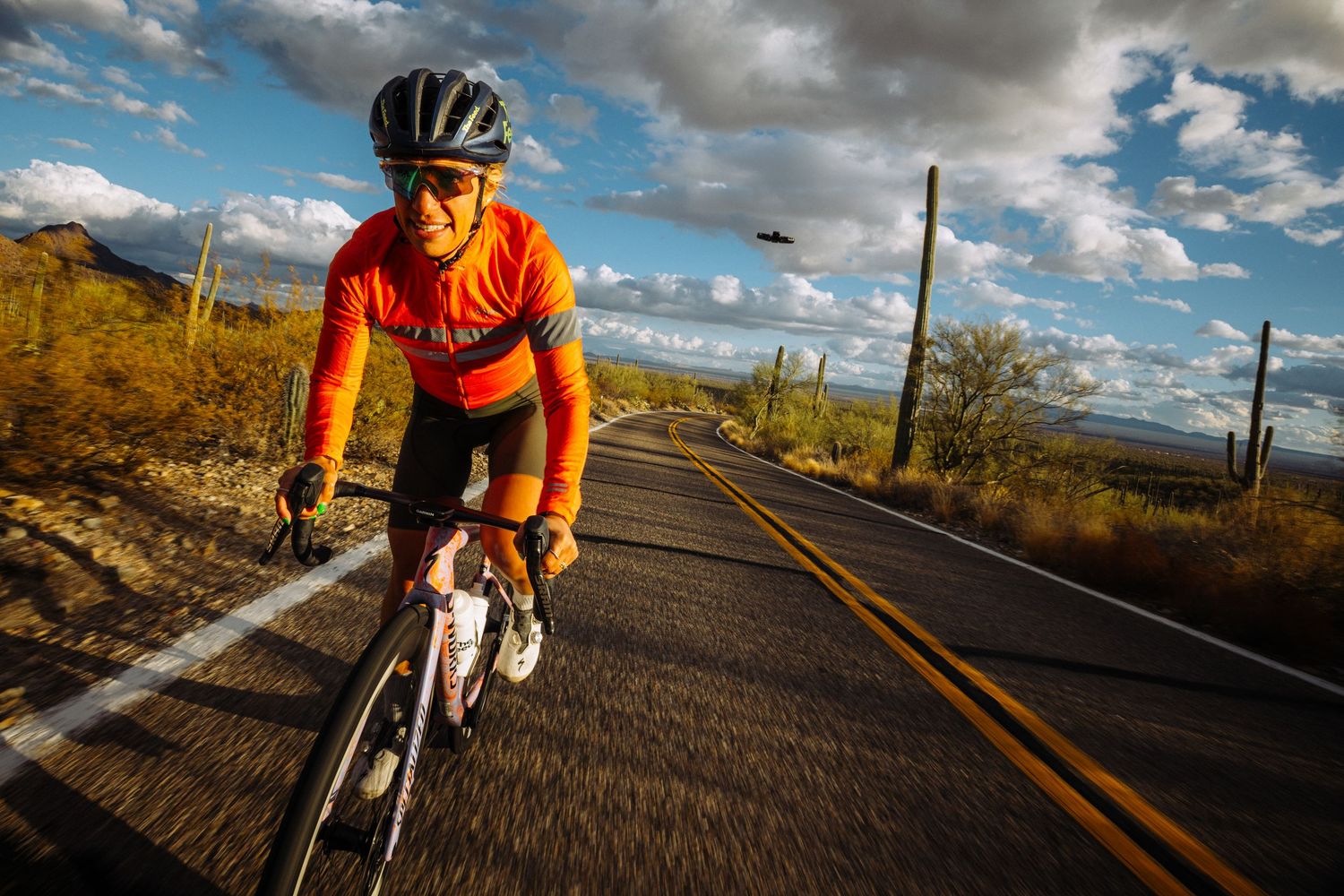 Cyclist in orange jersey riding road bike on desert highway with saguaro cacti and dramatic clouds at sunset.
