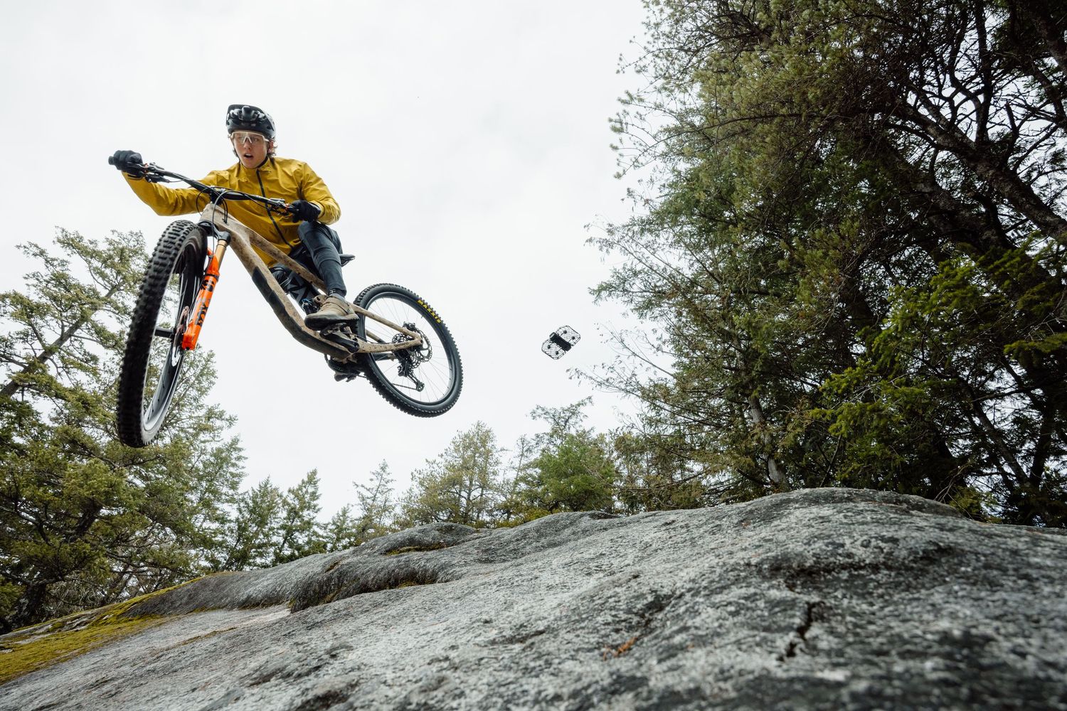 Mountain biker performs dramatic aerial jump off rocky terrain while wearing yellow jacket with drone flying nearby.