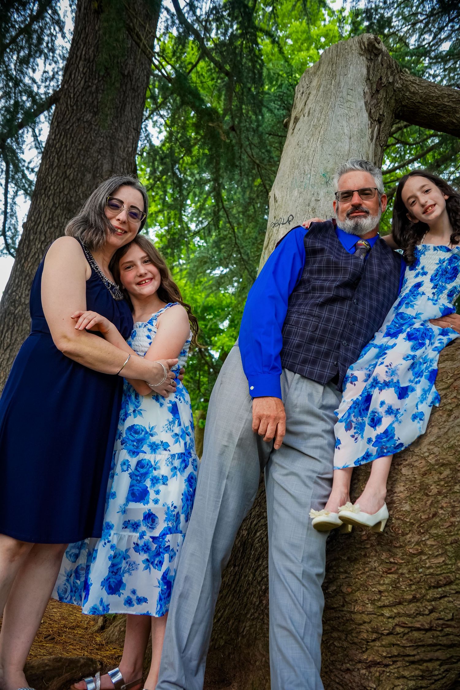 Family group poses creatively against large tree trunk in coordinated blue outfits.