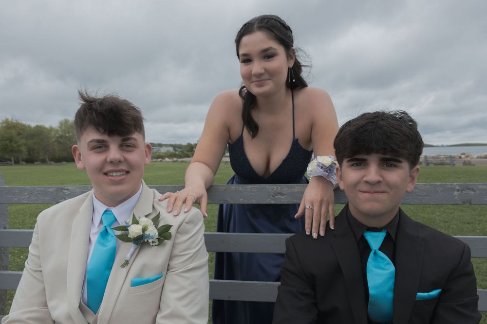 Three young people in formal attire pose together against a cloudy sky at an outdoor event.