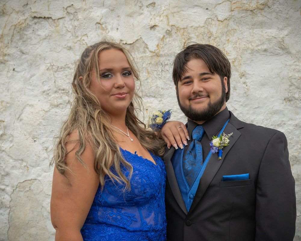 A couple in formal attire poses against a white stone wall at a formal event.