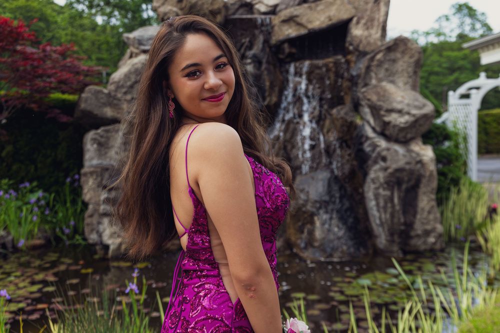 A figure in a sparkly purple dress poses by a rock waterfall feature in an outdoor garden setting.