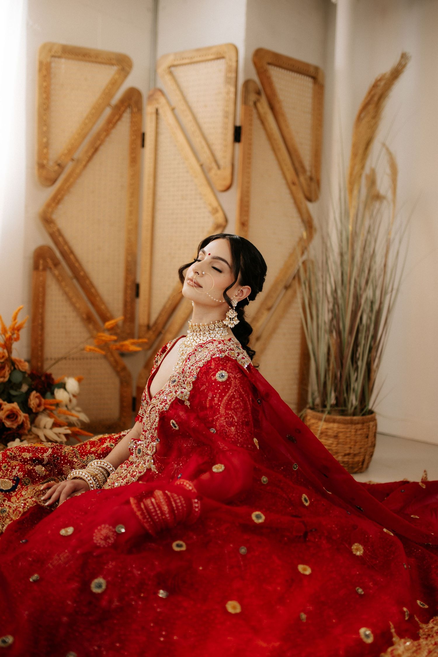 Elegant woman in vibrant red traditional lehenga with gold embroidery poses against a decorative folding screen backdrop.