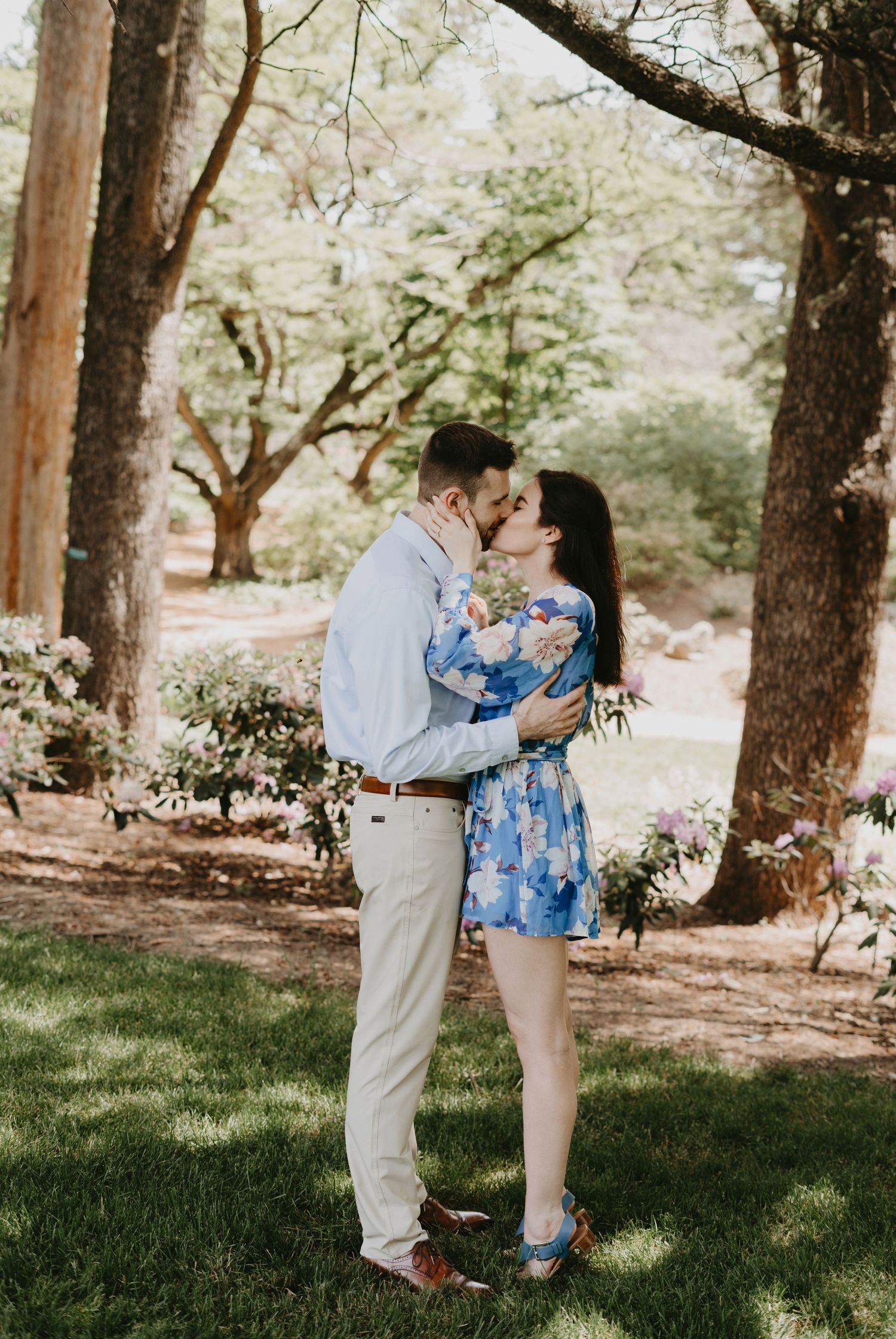 A tender embrace under trees in a park with spring flowers blooming.