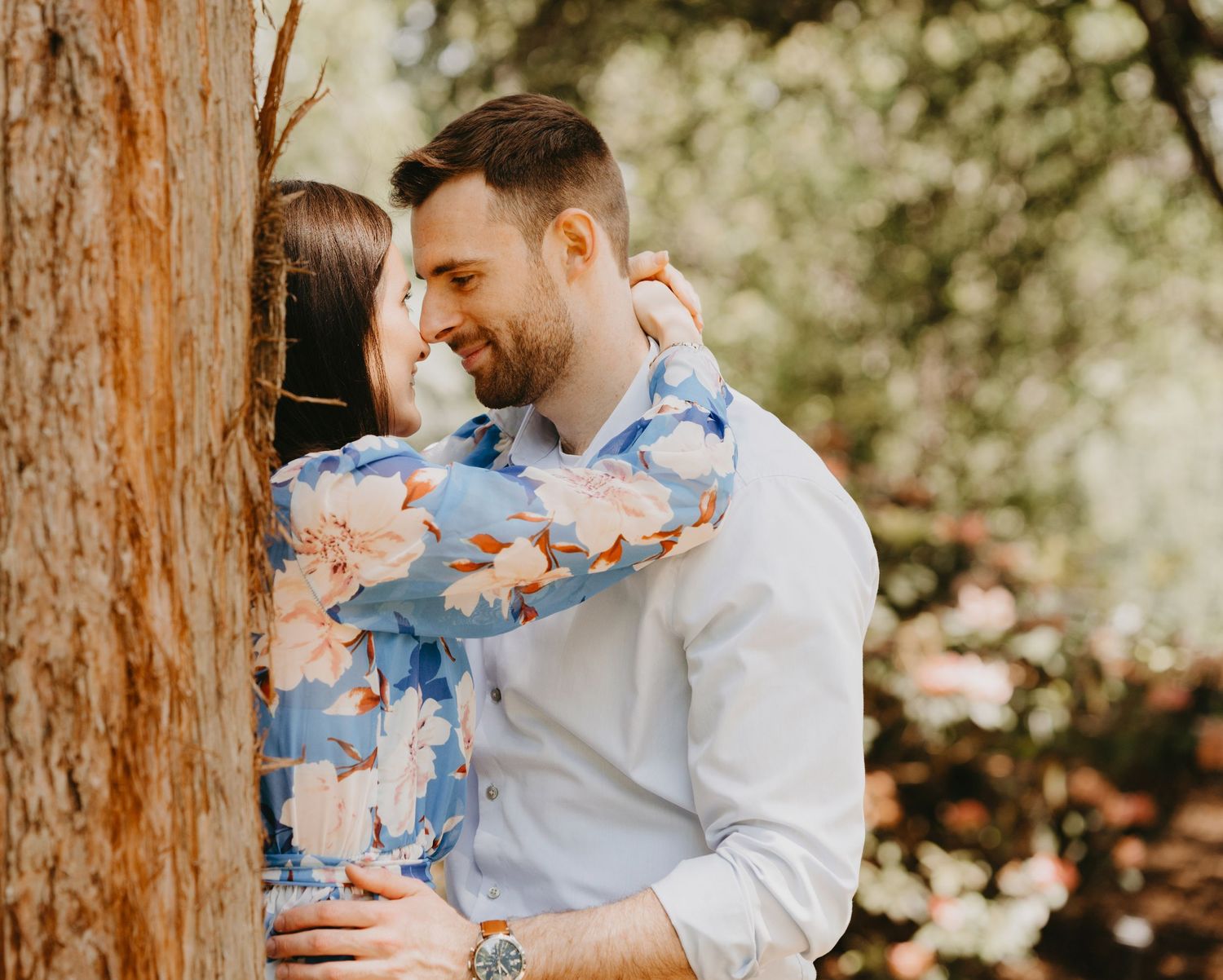 A loving couple shares an intimate moment against a large tree trunk.