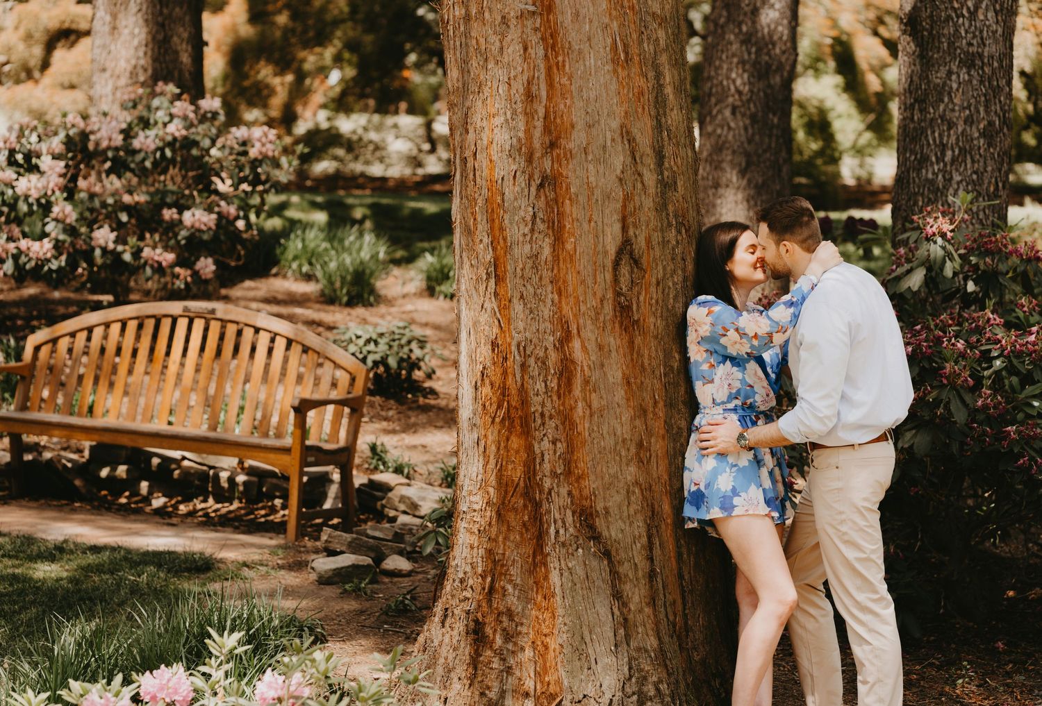 A romantic couple poses by a wooden bench and tree in a garden setting.
