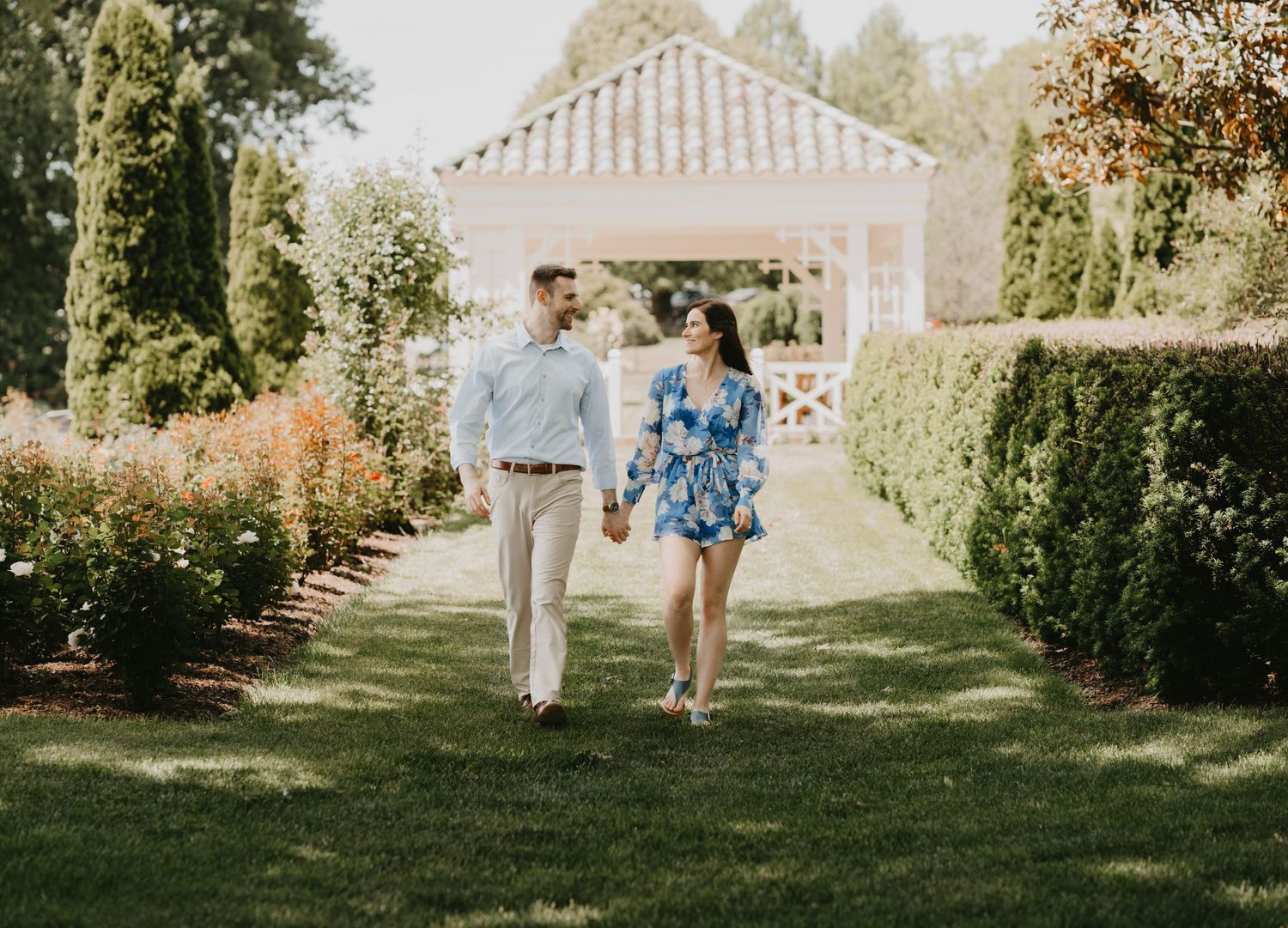 A couple walks hand in hand through a manicured garden path with white gazebo.
